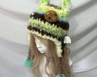 May include: A whimsical, handmade crochet hat in shades of brown, green, yellow, and blue. The hat features a cookie embellishment and fluffy ear-like extensions. Mint green pom-poms dangle from the sides, adding a playful touch to this unique accessory.