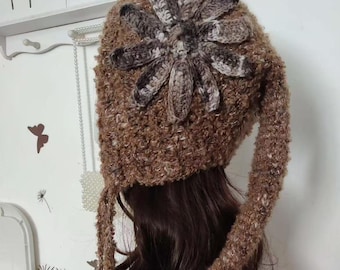 May include: A brown crocheted hat with a large flower design on top. The hat has long ear flaps and a textured appearance. The flower is made of a lighter brown yarn, and the hat is a darker brown color. The word "SWEETHOME" is visible in the background.