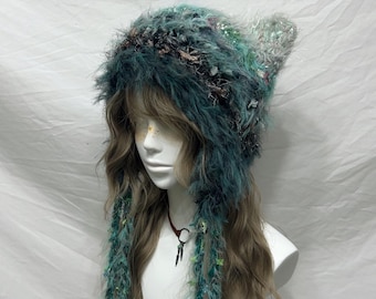 May include: A whimsical, handmade hat in shades of teal, green, and gray. The hat features long, fringed accents and a textured, furry design. The hat is displayed on a mannequin head with long, wavy brown hair.