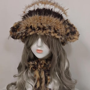 May include: A faux fur hat and scarf set. The hat features a wide brim and a layered design with brown, white, and tan fur. The scarf has a similar design with hanging fur accents. The set is displayed on a mannequin head with gray hair.