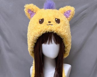 May include: A yellow plush hat with a cartoon bear face design. The hat features brown eyes, a small nose, and a purple tuft of fur on top. The ears and inner ear details are also purple.