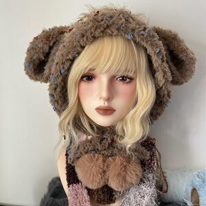 May include: A brown faux fur hat with large ear-like protrusions and a matching scarf. The hat and scarf are adorned with small blue and red accents. The scarf features two fluffy brown pom-poms and a patterned design with pink and purple hues.