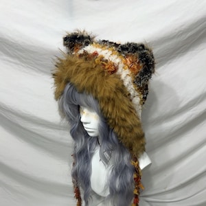 May include: A faux fur hat with a hood and ear-like accents. The hat features a mix of brown, white, and black fur, with orange and yellow accents. The hat is displayed on a mannequin head with long, wavy gray hair.