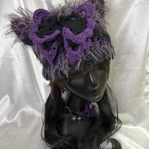 May include: A handmade hat with a large, crocheted purple and black butterfly design. The hat is primarily gray with fuzzy, textured accents. A matching purple flower adorns the neck of the mannequin.