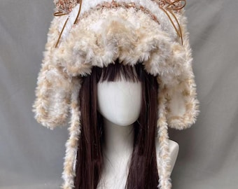 May include: A fluffy, faux fur hat in cream and tan colors. The hat has ear flaps and is decorated with brown accents and small bows. The hat is designed to be worn on the head and is displayed on a mannequin head with long, dark brown hair.