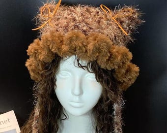 May include: A brown and tan fuzzy hat with a pom-pom trim and small, pointed ears tied with orange string. The hat is displayed on a white mannequin head with long, wavy brown hair. The background is black.