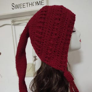 May include: A red crochet hat with long ear flaps and a tassel detail. The hat is made of a textured, knitted material and is displayed on a mannequin head. The word "SWEETHOME" is visible in the background.