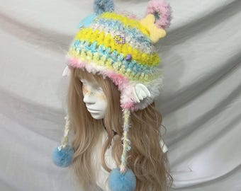 May include: A colorful, handmade crochet hat with a whimsical design. The hat features a mix of pastel colors, including blue, yellow, pink, and white. It has ear flaps, pom-poms, and decorative accents, creating a playful and unique accessory.