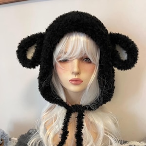 May include: A black, fluffy hat with bear ears. The hat is made of a soft, textured material and has a hood that frames the face. The ears are rounded and stand up from the top of the hat. The hat is tied with black strings.