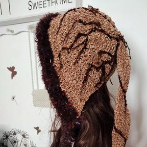 Könnte beinhalten: Ein brauner Häkelhut mit dunkelbraunem Besatz und Akzenten. Der Hut hat ein strukturiertes Design und eine lange, spitz zulaufende Klappe. Der Besatz ist dunkelbraun mit roten und violetten Tupfen. Ein einzigartiges Accessoire.