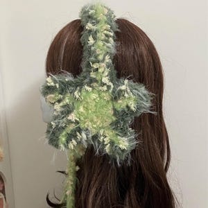 Op de afbeelding: Een handgemaakte, pluizige hoofdband in tinten groen, crème en grijs. De hoofdband heeft een stervormig ontwerp aan de zijkant, met een zacht, getextureerd uiterlijk. Het accessoire is ontworpen om op het hoofd te worden gedragen en voegt een unieke touch toe aan elke outfit.
