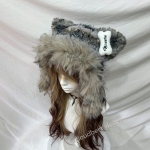 Puede incluir: Un sombrero de piel sintética con orejas adjuntas y un parche en forma de hueso que dice "Puppy". El sombrero es principalmente gris y marrón, con una textura esponjosa. El sombrero está diseñado para parecerse a un lobo o un perro.