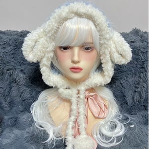 May include: A fluffy white and light blue hat with bunny ears, worn by a mannequin. The hat has a soft, textured appearance and is tied with a pink ribbon. The mannequin has white hair and is wearing the hat.