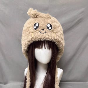 May include: A tan, fluffy hat with a cartoon face design. The hat features large, black-outlined eyes and a smiling mouth. The hat has ear flaps and a small, rounded top. The hat is designed to be worn on the head.