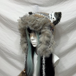 May include: A faux fur hat with a hood and ear flaps. The hat is primarily gray and brown with a white accent on the ear. The hat is on a mannequin head with blue and black hair.