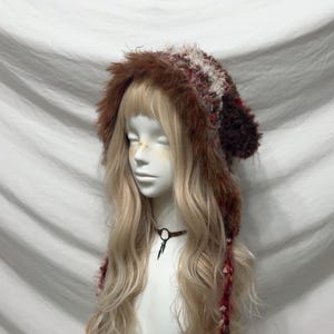 May include: A brown and red faux fur hat with ear flaps and a textured design. The hat is displayed on a mannequin with long blonde hair and a brown choker necklace.