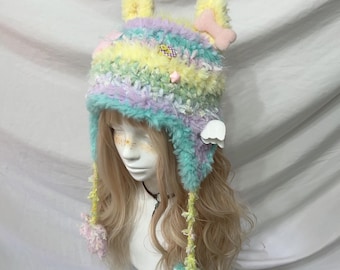 May include: A colorful, fluffy hat with a whimsical design. The hat features a mix of pastel colors, including yellow, blue, and purple. It has ear-like protrusions, a bone-shaped accent, and dangling pom-poms. The hat is designed to be worn.