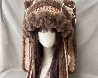 May include: A brown faux fur hat with attached ears and long, braided accents. The hat features a mix of brown and beige tones, with small white bows at the top. The hat is designed to be worn on the head.