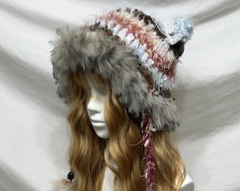 May include: A unique, handmade hat with a faux fur trim and a colorful, textured crown. The hat features a mix of pink, blue, and brown yarns, with dangling beaded accents and fluffy pom-poms. The hat is designed to be worn on the head.