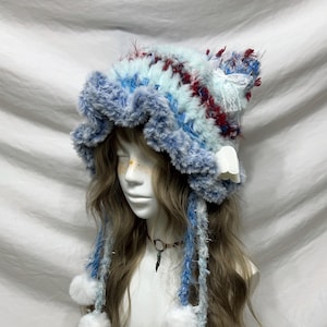 May include: A whimsical, handmade hat in shades of blue, white, and burgundy. The hat features a fluffy, textured brim and decorative accents, including dangling strands with pom-poms and a small, white, wing-shaped embellishment. The hat is designed with a unique, artistic flair.