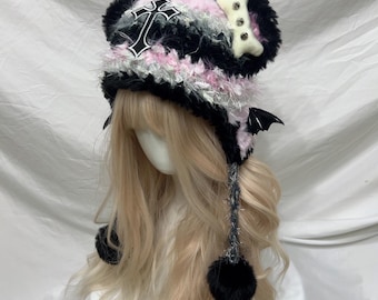 May include: A unique, handmade hat with a gothic aesthetic. The hat features a black and white cross, a bone-shaped embellishment, and bat wings. It is made with a combination of black, pink, and gray fluffy yarn, and has black pom-poms and dangling accents.