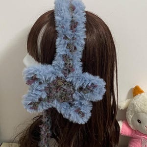 Könnte beinhalten: Ein flauschiges, hellblaues Stirnband mit einem sternförmigen Akzent. Der Stern hat violette, grüne und braune Akzente. Das Stirnband besteht aus einem weichen, flauschigen Material. Ein Plüschlamm-Spielzeug ist im Hintergrund zu sehen.