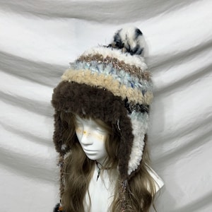 May include: A handmade winter hat with ear flaps and a pom-pom. The hat features a mix of textures and colours, including brown, white, beige, and blue. The ear flaps are trimmed with a dark brown, fluffy material.