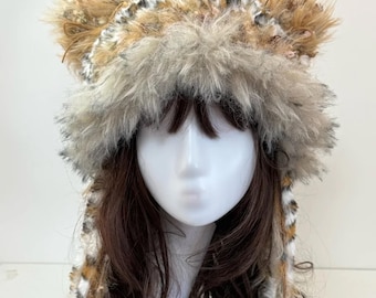 May include: A faux fur hat with attached ears, featuring a mix of brown, tan, and cream colors. The hat has a fluffy texture and is designed to resemble a wild animal. The hat is displayed on a mannequin head.
