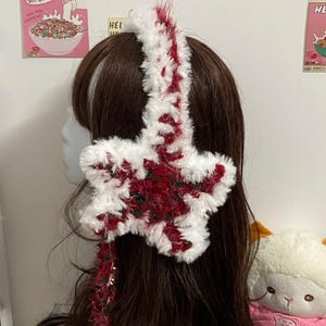 Pode incluir: Um acessório de cabelo em forma de estrela fofa branca e vermelha. A estrela é decorada com detalhes vermelhos e verdes. O acessório de cabelo é usado em cabelos castanhos. O tema geral é festivo e decorativo.