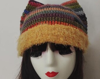 May include: A handmade, colorful crochet hat with cat ears. The hat features a mix of rainbow colors and a fuzzy, yellow brim. The hat is displayed on a mannequin head with dark hair, long eyelashes, and red lipstick.