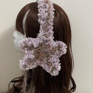 Op de afbeelding: Een zachte, lavendelkleurige haarband met een grote, vijfhoekige ster. De ster en haarband zijn gemaakt van een zacht, getextureerd materiaal. Het accessoire wordt getoond op een mannequinhoofd met bruin haar.