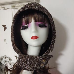 Puede incluir: Una capucha de punto marrón y blanco con cierre de botón. La capucha está diseñada para cubrir la cabeza y el cuello, brindando calidez y estilo. La palabra "SWEETHOME" es visible en el fondo.