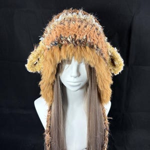 May include: A handmade, furry hat with ear flaps and long braided ties. The hat features a mix of brown, tan, and cream-colored faux fur and yarn. The hat is displayed on a mannequin head against a black background.