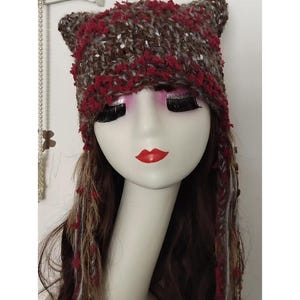May include: A handmade cat ear hat in shades of brown, red, and white. The hat is made of a textured yarn and features pointed ears. The hat is displayed on a mannequin head with long brown hair and red lipstick.