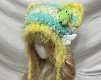 May include: A whimsical, handmade hat in shades of yellow, blue, and white. The hat features a fluffy texture, a small green plush frog, and white wing accents. The hat has long, dangling ties with pom-poms.