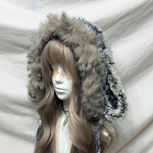 May include: A furry, hooded hat with long, floppy ears. The hat is primarily gray with black accents and a fluffy texture. The hood frames a face with long, wavy, light brown hair. The hat has a whimsical, fantasy-inspired design.