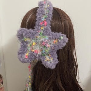 Op de afbeelding: Een pluizige, lavendelkleurige ster-vormige haarband met kleurrijke draadaccenten. De ster is bevestigd aan een bijpassende lavendelkleurige band. De haarband wordt op bruin haar gedragen.