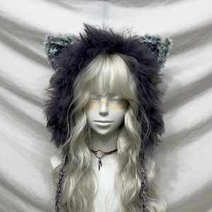 Puede incluir: Una capucha de piel sintética gris oscuro con detalles de orejas a juego. La capucha se usa sobre una peluca larga, ondulada y rubia clara. Se ve un collar pequeño y oscuro con un colgante.