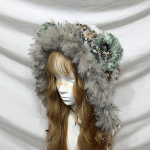 May include: A hooded head covering with a faux fur trim in shades of grey and green. The hood is adorned with various textures and embellishments. The head covering is displayed on a mannequin head with long, wavy, light brown hair.