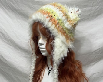 May include: A handmade, colorful crochet hat with ear flaps. The hat features a mix of yellow, orange, and white yarn, with fluffy white trim and yellow pom-poms. The hat is designed to be worn in cold weather.