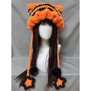 May include: A handmade orange and black tiger-striped hat with long, dangling black and orange braids. The hat features fluffy orange trim and black pom-poms and star-shaped accents at the ends of the braids. The hat is displayed on a mannequin head.