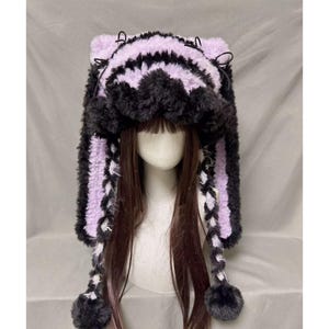 May include: A fluffy, black and lavender hat with ear-like protrusions and braided accents. The hat features a striped pattern on the top and sides, with small pom-poms at the end of the braids. The hat is designed to be worn on the head.