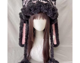 May include: A plush, black and pink hat with ear flaps and paw-shaped accents. The hat features a striped pattern on the top and pink accents on the inside of the ear flaps. The hat is designed to be worn on the head.