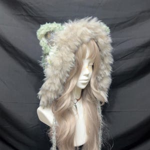 May include: A faux fur hood hat in shades of grey, beige, and green, displayed on a mannequin head with long blonde hair. The hat features a fluffy texture and ear-like details, with long, dangling strands.