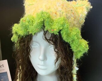 May include: A handmade, fluffy hat in shades of yellow and green, with a ruffled brim. The hat is made of a textured yarn and has long, thin ribbons. The hat is displayed on a white mannequin head with long, brown curly hair.