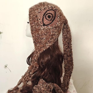 Può includere: Un cappello all'uncinetto marrone e beige con un lungo orecchio pendente e una sciarpa abbinata. Il cappello presenta un disegno circolare al centro. Il cappello e la sciarpa sono realizzati con un filato strutturato.