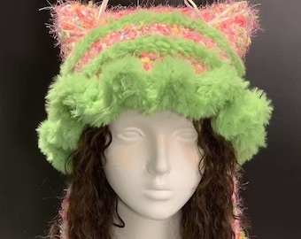 May include: A whimsical cat ear hat in shades of pink, green, and yellow. The hat features a fluffy green brim and cat ears, with a striped pattern of pink and green. The hat is made of a soft, textured material.