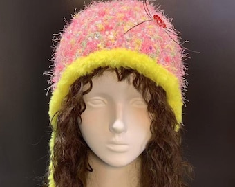 May include: A pink and yellow knit hat with a fuzzy texture. The hat has a yellow brim and a small red embellishment on top. The hat is displayed on a mannequin head with brown curly hair.