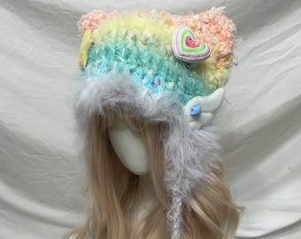 May include: A colorful, handmade hat with a rainbow design. The hat features a fluffy, textured material with accents of pink, yellow, blue, and green. It is adorned with a heart-shaped embellishment and small white wings.