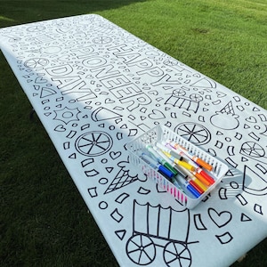 May include: A white rectangular tablecloth featuring black line art patterns of covered wagons, ice cream cones, and hearts with the text HAPPY PIONEER DAY. A white plastic basket filled with assorted colorful markers sits on the table surface.